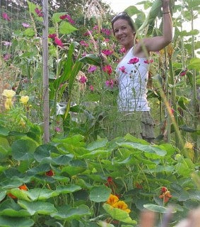 Woman in a white top stands among green leaves and flowers that are pink, yellow, and orange