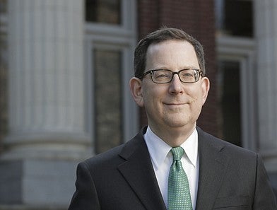 A man with glasses, a green tie, white shirt and grey jacket smiles