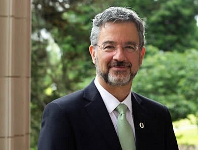 A man with gray hair, moustache beard, glasses, a light green tie, white shirt, and gray jacket smiles