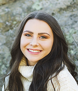 Young white woman with long, dark brown hair and wearing a beige sweater smiles broadly at the camera