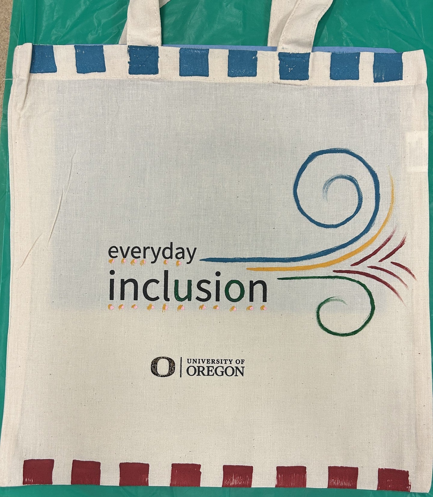 Everyday Inclusion tote bag with blue boxes on top and red boxes on the bottom. U and O of Everyday Inclusion colored green. Blue, yellow, green, red swirls at right