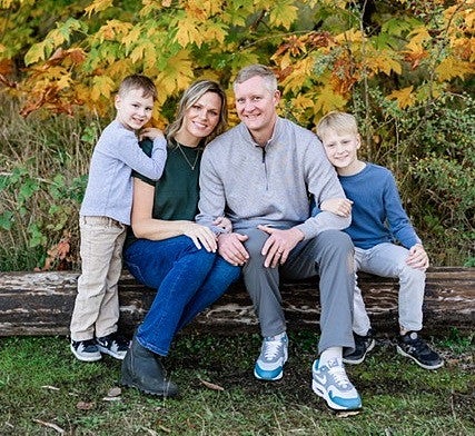Ashley sitting on a log outside; one son stands next to her at left; her husband seated next to her at right; their other son seated on the log on the far right