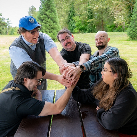 Five people around a table with their hands together struggling against one woman whose hand is at the centerstruggling 
