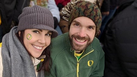Woman in a grey woolly hat and scarf with a UO "O" on her cheek next to a man with a beard and green UO "O" top.