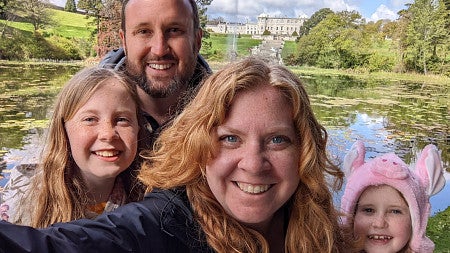 Smiling woman with long red hair; behind her are two young girls and a white man with dark hair, moustache and beard; green fields and trees in the background with a castle and blue skies