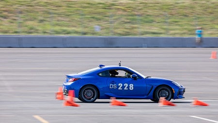 Blue racing car with DS228 on the side; a person in a helmet visible in the car; 4 orange traffic cones on their side; blurry because the car is in motion
