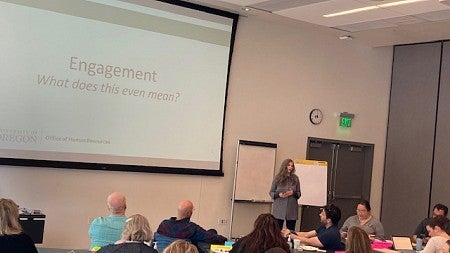 Woman with long sandy hair and a gray jacket addresses a seated group; slide reads Engagement - What does this even mean?