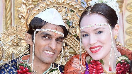 Young Indian man at left and young white woman at right; both have dark hair; wearing traditional Marathi wedding attire including red petal garlands; gold ornament wall behind them