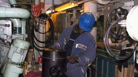 African man in a blue helmet and blue overalls with his head down focusing on his work. It looks like he is in the engine room of a ship.