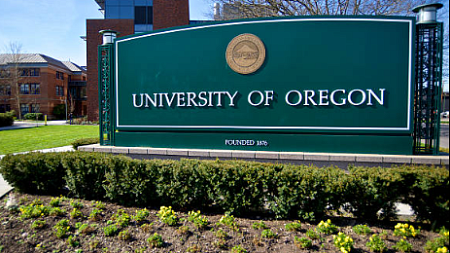 Green sign with University of Oregon and Founded 1876 in white; green bushes in front; green grass and red buildings in background