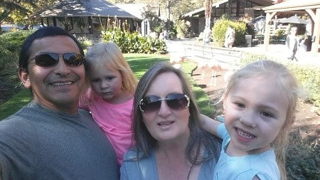 Man with short black hair and sunglasses holding his daughter; next to him is a woman with long brown hair and sunglasses holding their other daughter