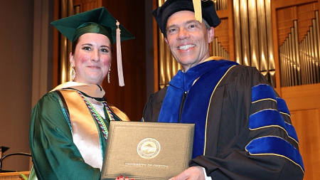 Elisabeth in green and gold graduation gear receiving her MBA from Bruce Blonigen (white man, dean of the business college)