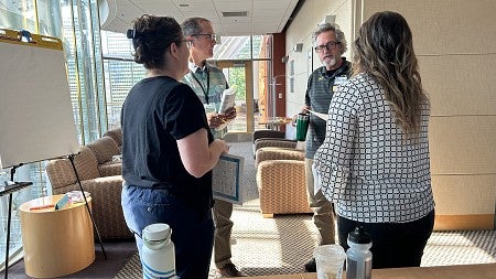 Two men and two women standing talking with papers in their hands; sunlight streams in windows to their left