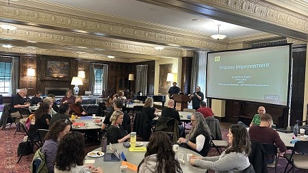 Groups of 6 people seated around tables; Deborah Butler at the front of the room speaking on a microphone; Process Improvement slide projected behind her