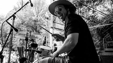 Black and white image of a man in t-shirt, shorts, hat, and sunglasses playing drums on a stage; microphones and audience visible in front of him