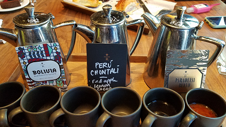 Three silver coffee pots, each holding different coffee arranged on a tray behind and six grey cups.