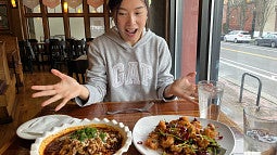 Woman with a knitted cap and grey GAP sweatshirt sits at a table with hands open in amazement and joy; she is looking at two dishes of delicious Szechuan Chinese food; two glasses of water are on the table; a street is visible through the window to the right