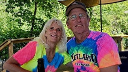White woman with blonde hair and colorful Tshirt at left; at right a white bespectacled man with colorful T-shirt and ball cap; green trees in background