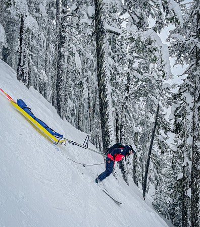 Figure in patrol uniform and helmet on skiis with a sled behind--skiing down a steep snowy downhill; snow covered trees behind