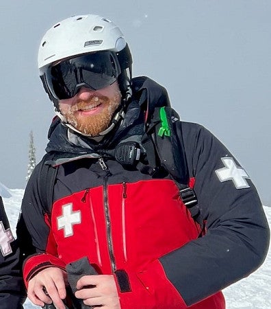 White man with ginger beard and moustache, black goggles, white helmet, and red-blue uniform on a snowy hill with snow sky behind