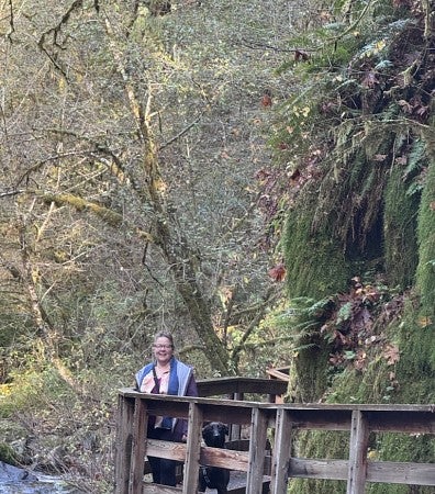 White woman with brown hair stands on a wooden walkway in the woods with her black dog. Moss-covered rocks, trees, and a river in the background.