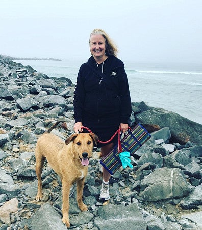White woman with long blonde hair stands next to a big brown dog (Shaggy) on rocks in front of the sea