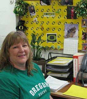 Dee at work: woman in a green Oregon Ducks T-shirt smiles; behind her are a lot of yellow and green UO decorations 