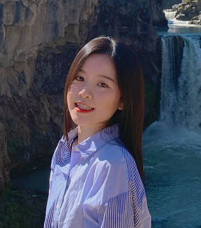 Young woman with long dark hair, wearing a light blue collared shirt smiles with a waterfall and river in the background