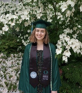 Young woman with shoulder-length brown hair, in UO College of Design graduation wear, smiles in front of white flowers outside