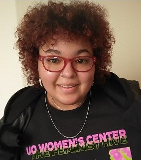 Young woman with red glasses and shoulder length brown hair smiles at the camera; she wears a black shirt with the text: UO Women's Center, The Feminist HIve
