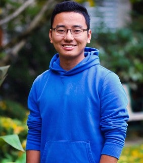 Young man with short black hair and glasses, wearing a blue hoodie, smiles. Greenery visible in the background.