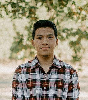 Young man with short black hair, wearing a red, white, and black checkered, collared shirt stands outside with trees and sunlight in the background