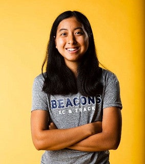 Woman with long black hair and folded arms, wearing black pants and a grey t-shirt that reads "Beacons XC & track", smiles in front of a yellow background