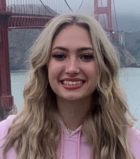 Woman with long blonde hair and a pink sweatshirt stands in front of the Golden Gate bridge and bay on a misty day