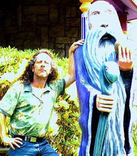 Man with long brown hair, floral shirt, jeans, and hand on his hip leans on the shoulder of a statue--of a sage with a long grey beard and staff. Flowers visible behind the man.