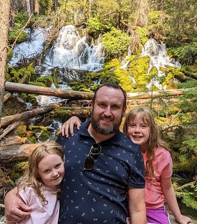 White man with moustache and beard between to his two daughters (both with blonde hair) standing in front of logs, trees, and a waterfall