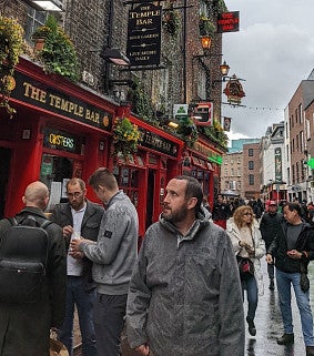 Man in a grey coat looks up at a bar; behind him is The Temple Bar, lots of people walking in the street; grey skies and stone buildings