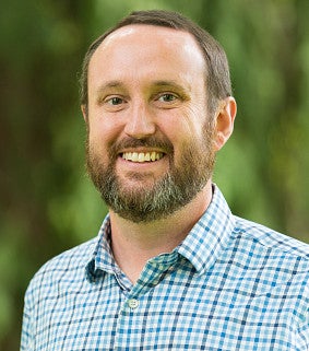 Smiling white man with brown hair, beard and mustache; wearing a checkered blue-white collared shirt; greenery blurred in the background