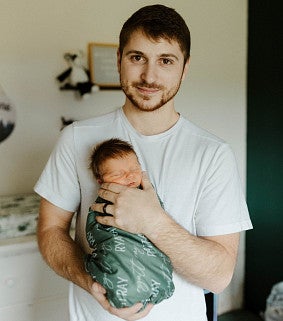 Young white man with short dark hair, moustache, and short beard holds a small baby wrapped in green