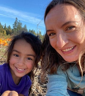 Young girl with brown hair and purple shirt; woman with brown hair and blue shirt; pumpkin field, trees, and blue sky in the background.