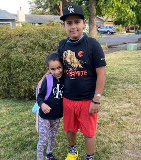 11-year old boy in a ball cap, black T-shirt and red shorts stands with his arm around his 6-year-old sister in a sweatshirt and long pants; backpack over one shoulder; standing on grass