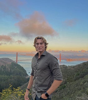 Young white man with wavy blond hair wearing a grey collared shirt standing in front of green hills, a bay, and the Golden Gate bridge in the distance