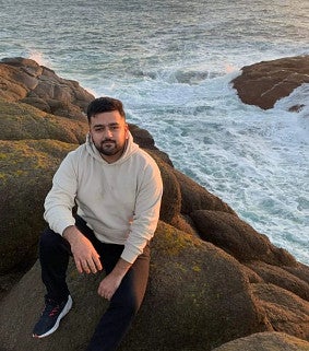 Young man with short dark hair, moustache and beard sitting on the rocks resting his right arm on his folded knee. White top and dark trousers. Crashing waves and ocean behind him.