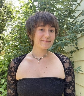 Young white woman with short brown hair, lacy black top and necklace stands in front of leafy green foliage outside