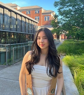 Asian woman with long black hair, a white dress and beige graduation stole stands outside with green grass, trees and buildings behind her