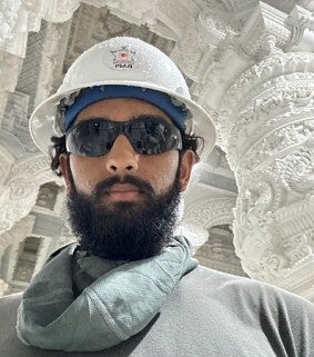 Asian man with moustache and beard wearing sunglasses and a hard hat looks at the camera; ornate white temple ceiling visible in the background