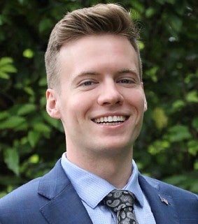 Young whilte male with short hair, blue jacket, blue shirt, and tie smiles into the camera; greenery behind him