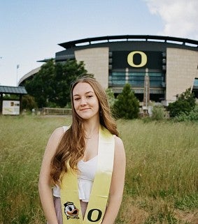 Young white woman with long brown hair, white dress, and gold graduation stole stands in front of Autzen Stadium; UOregon O is visible on the stadium; light blue sky with clouds