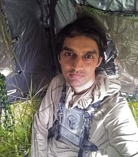 Young Indian man with black haer and wearing a grey hoodie. Green grass and a tent-like structure behind him. 