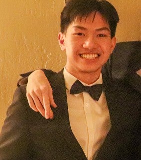 Young East Asian man in a tuxedo and bow tie stands and smiles at the camera; a hand draped over his right shoulder; beige wall in background